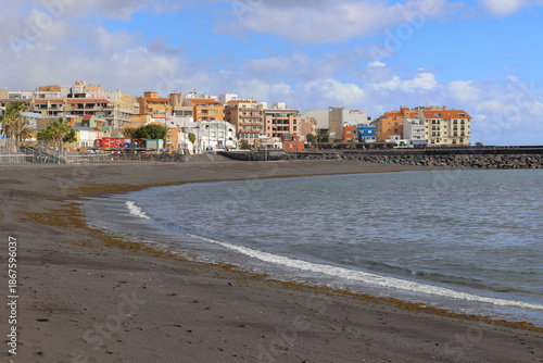 Horizontal seaside landscape showing a curved black sand beach with gentle waves and a rocky breakwater. 