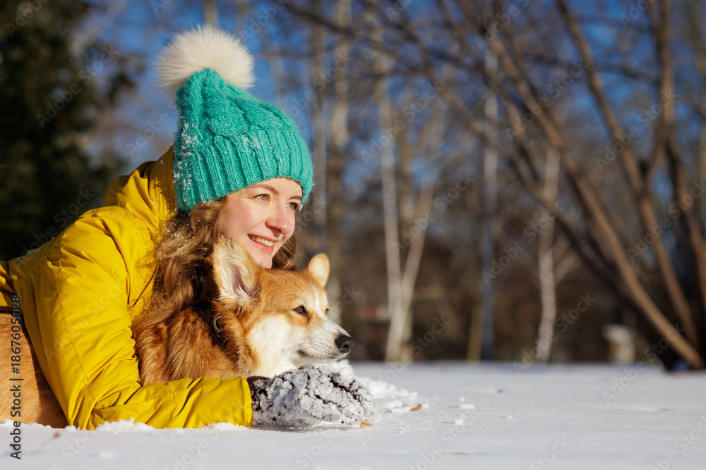 Fototapeta premium Woman hugging corgi on snow in winter park with copy space on sunny day