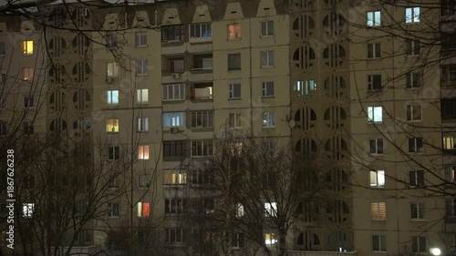 Quiet night scene of a vintage residential building with softly glowing windows, leafless trees framing the facade, capturing calm urban solitude under gentle night lighting
