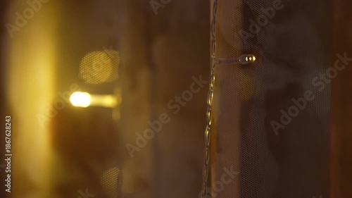 Night winter city view through window with warm glowing lights on curtains, apartment building outside, cozy home interior atmosphere and urban lifestyle concept