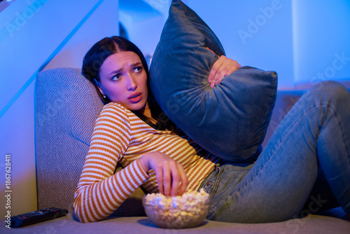 Quadro em tela Young scared woman watching horror movie eating popcorn and hugging pillow, sitt