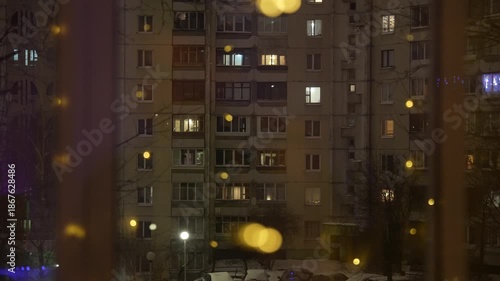 Night winter city view through window with warm glowing lights on curtains, apartment building outside, cozy home interior atmosphere and urban lifestyle concept