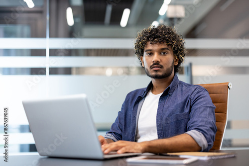 Foto Portrait of a young Indian man working in an office, sitting at a desk with a la