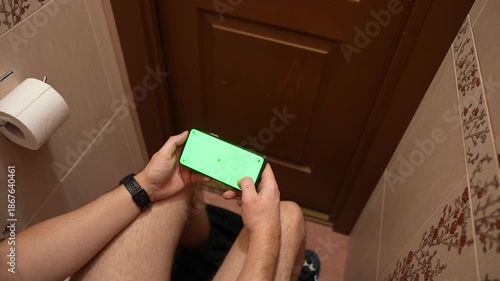Top view of man using smartphone with green screen while sitting in restroom, hands scrolling on mobile phone, casual daily routine, technology mockup and modern lifestyle concept