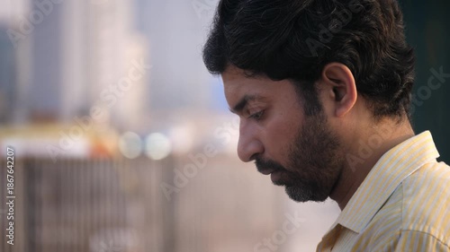An outdoor daylight close up shot of a middle aged man or male looking at or glued to the screen of a mobile phone with blurred vehicles passing on a busy city road and urban skyline in the background