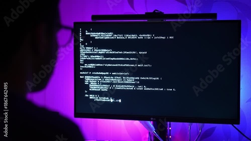 Over-the-shoulder or back view of a man working on a computer with code on the monitor. Face is hidden in shadow, only glasses slightly visible. The scene is illuminated with purple and blue RGB light