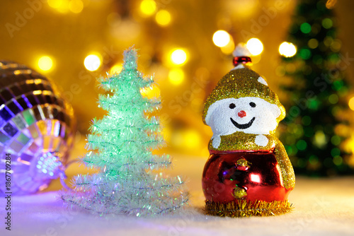 On a blurred background with bright yellow lights are two Christmas trees with a large ball and a snowman
