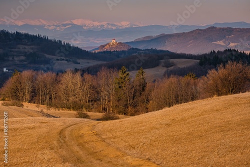 Peaceful hilly countryside landscape with a small church on a hill, winding dirt path and forested hills. Distant mountain range and soft light create a calm rural atmosphere.