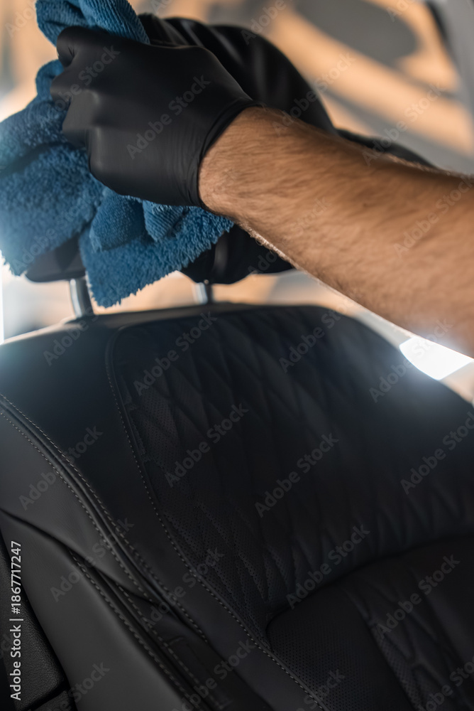 Obraz premium A person in black nitrile gloves wipes a car seat headrest with a blue microfiber towel inside a vehicle. Quilted perforated leather and contrast stitching show premium care.