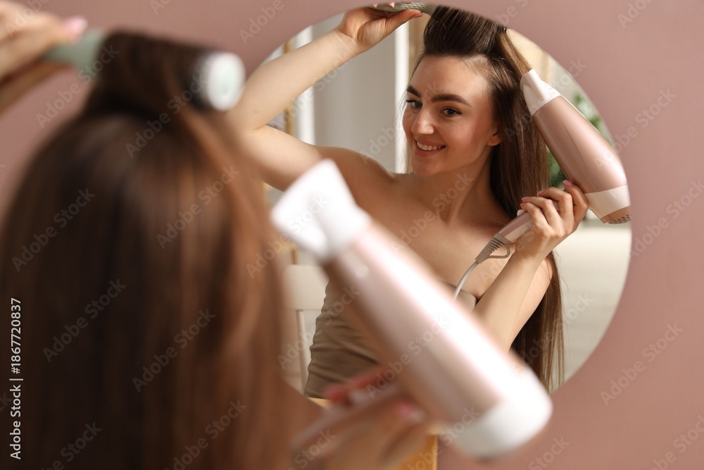 Fototapeta premium Smiling woman styling her hair with hairdryer and brush near mirror indoors