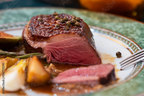 A cut piece of duck breast meat topped with green peppers in a plate with its original orange sauce with side dish vegetables.