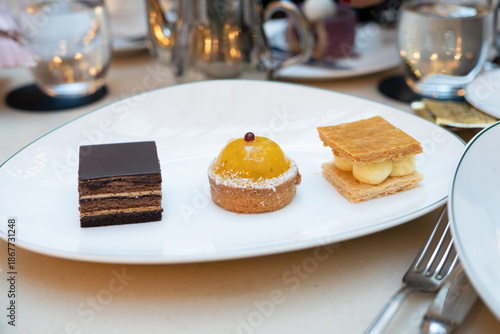 A piece of assorted mini french dessert such as mango tart, traditional opéra pastry and millefeuille in a white plate with fork and cup of tea in background 