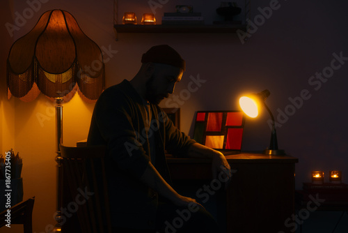 Atmospheric interior scene of a man sitting at a desk illuminated by warm lamps and candlelight, creating a calm and introspective mood. The image highlights cozy home ambiance and cinematic lighting.