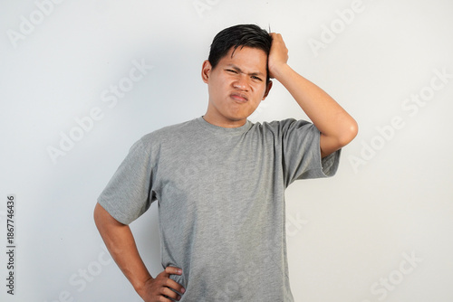A young Asian man in a gray shirt with his hand on his head, showing confusion and indecision.