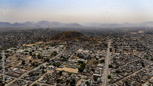 Aerial view of a sprawling cityscape nestled beneath a majestic mountain range, roads stretching like arteries through the urban landscape, Jalingo, Taraba, Nigeria.