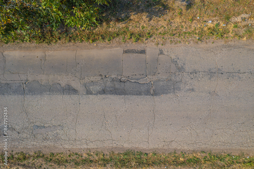 Obraz premium Cracked asphalt road surface with damaged patches and dry roadside grass