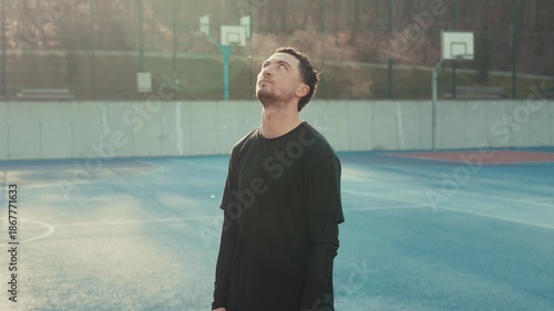 A serious young man in a black tee stands on a sun-drenched outdoor court. Intense gaze and focused expression under a warm direct sun light light. Pissed expression followed by a facepalm.