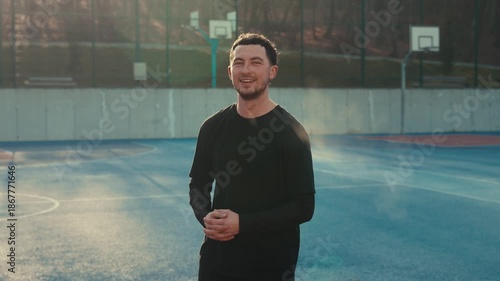 A cheerful brunette young man in a black shirt smiles and turns to the camera on a bright colored outdoor court. direct sunlight creates a warm glow on his face and hair.
