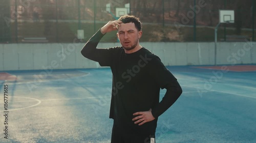 Pensive young brunette basketball player on a vibrant blue court, with his hand to his head in deep concentration. Direct sunlight from the behind. Black sportswear