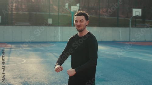 A young man playfully flexes his muscles on a vibrant blue basketball court. Golden hour backlight creates a warm, cinematic rim light. Energetic, triumphant, and urban atmosphere with a gritty, sun-d