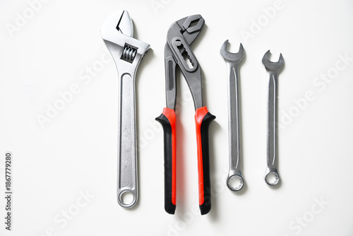 Top view of an adjustable wrench, water pump pliers with red handles, and two combination spanners arranged on a white surface. Hand tools for repair, maintenance, and DIY projects.