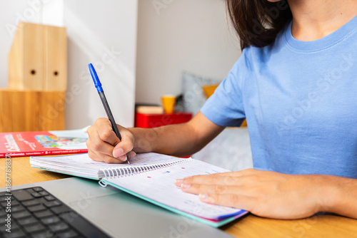 Young student girl writing notes in her notebook while studying at home, with a laptop and textbooks nearby