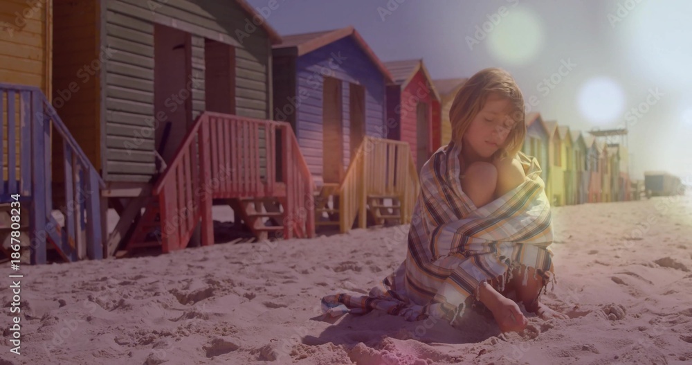 Fototapeta premium Sitting child wrapped in striped blanket digging in sand at beach, with colorful huts, copy space