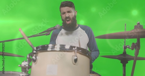 Singing bearded drummer playing drum kit in green screen studio, wearing raglan shirt with mic
