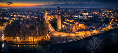 Aerial panorama of Casale sul Sile at Christmas sunset. Festive lights along the Sile river, illuminated historic tower and townscape.