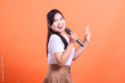 A cheerful young woman sings with a microphone, smiling and looking at the camera, raising her hand and enjoying her performance isolated on orange background