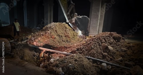 Excavator digging into a large pile of dirt at a construction site in front of a building at night.