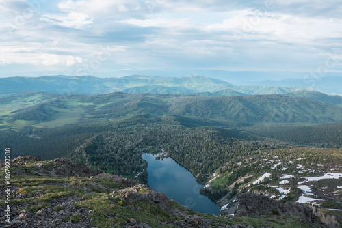 Top view from rocky precipice edge to blue lake in woody hilly terrain against mountain range silhouette far away. Cloudy sky reflected in smooth water surface of long forest lake among green hills.
