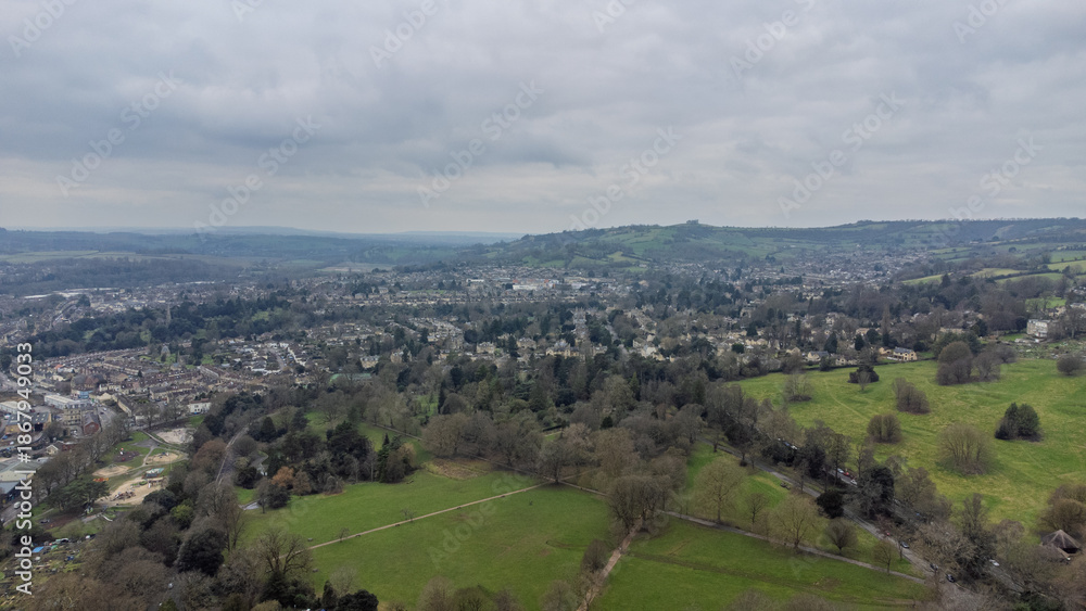 Fototapeta premium High aerial view over Bath showing a large park with intersecting paths, scattered trees, surrounding neighbourhoods of historic houses, and rolling green hills under an overcast sky.