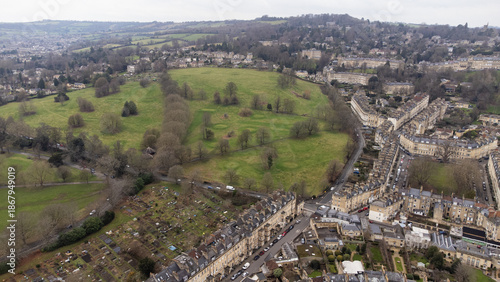 Aerial view of Bath showing a large park bordered by Georgian houses, curving streets, gardens, and dense historic housing set against surrounding hills.