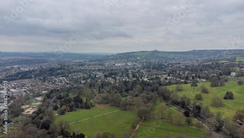 High aerial view over Bath showing a large park with intersecting paths, scattered trees, surrounding neighbourhoods of historic houses, and rolling green hills under an overcast sky.
