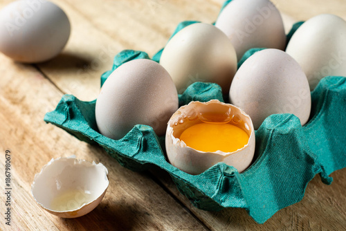 white eggs inside the carton, one egg broken in half with the shell on the wooden table