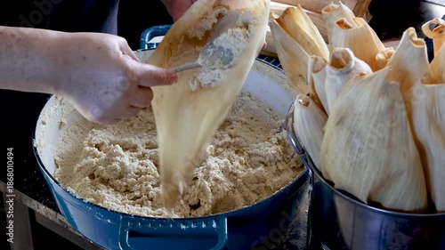 Woman's hands spreading masa dough on a corn husk to make traditional tamales in a rustic kitchen