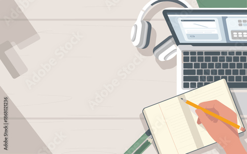 A person is writing in a notebook with a pencil beside a laptop and headphones on a wooden desk viewed from above.