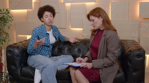Worried attractive African American woman sitting on sofa, telling and sharing about mental problems while attentive middle aged female counselor making notes during psychotherapy session.