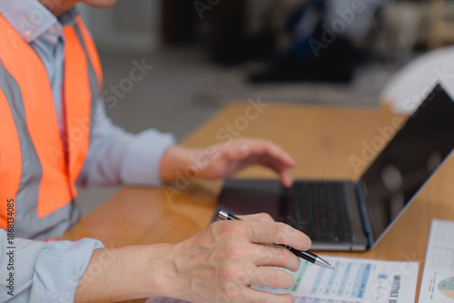 Engineer or inspector wearing safety vest checking documents while using a laptop at a construction site. Concept of industrial work, project management, inspection, and safety compliance.