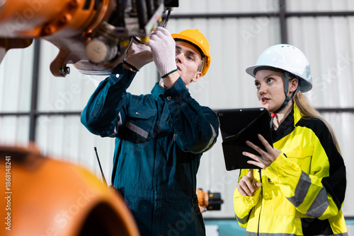 Male and female engineers in safety uniforms collaborate using digital tablets to control robotic arm equipment in smart factory. Concept of teamwork, automation and industry innovation.