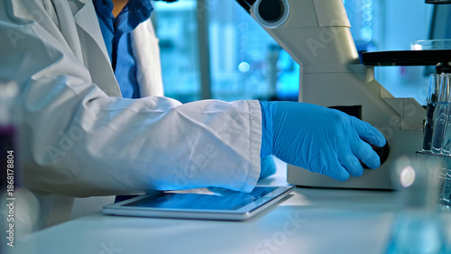 Vászonkép A scientist in a lab coat and blue gloves operates a microscope, analyzing samples in a well-lit lab with scientific equipment