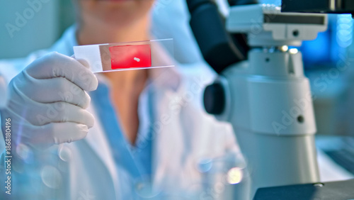 A scientist in a lab coat and gloves examines a red blood smear under a microscope in a clinical lab, highlighting precision in medical research and diagnostics.