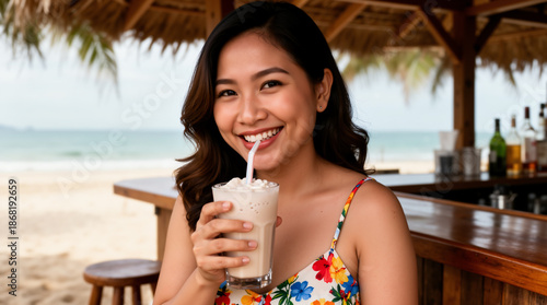 A cheerful Filipina woman with wavy dark hair smiles brightly while enjoying a creamy milkshake at a tropical beach bar, wearing a vibrant floral sundress against a coastal backdrop.