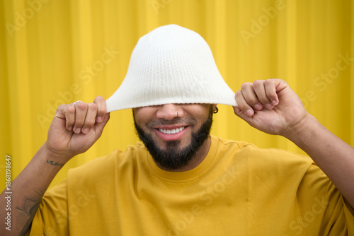 Smiling man wearing white hat playfully pulled over eyes against yellow wall