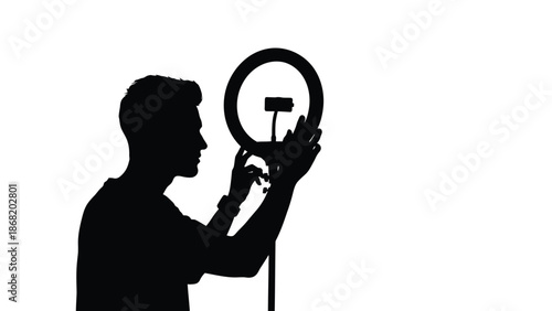 Silhouette of a male content creator adjusting a professional ring light and phone mount in preparation for recording video or live streaming against a stark white background isolation
