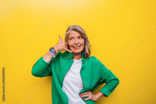 Mature woman in green jacket making call me gesture on yellow background