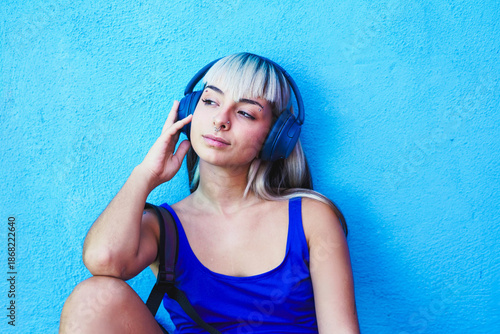 Woman in blue tank top with headphones listening to music by blue wall