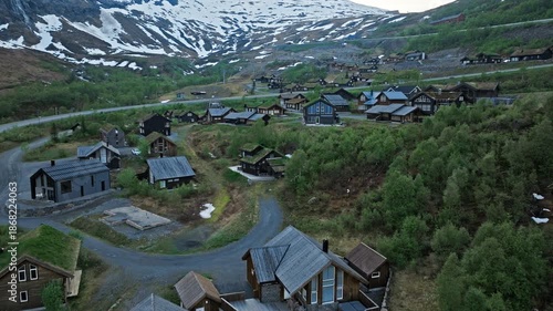 Aerial view of mountain houses at Røldal Skisenter in Norway. Drone footage showing alpine houses, winding roads, and green hills with snow patches during a calm summer evening.