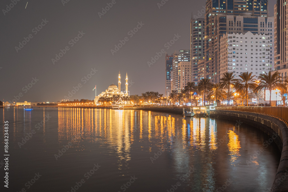 Obraz premium Sharjah, UAE,10.10.2025. A mosque stands in the distance, along a bustling waterfront with palm trees.